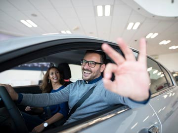 Eine Person sitzt auf dem Fahrersitz eines Autos und macht mit der Handgestik das 'OK'-Zeichen, während ein Beifahrer neben ihr in einer hellen, Innenumgebung sitzt.