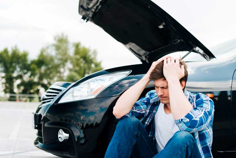 Ein Mann sitzt auf dem Boden neben einem schwarzen Auto mit angehobener Motorhaube und zeigt Anzeichen von Stress und Frustration.